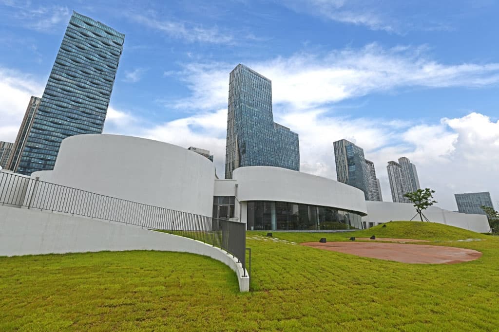 잔디밭 위 흰색 곡면 벽으로 이루어진 국립세계문자박물관 외관과 뒤편 송도의 고층 빌딩